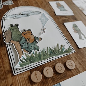 May include: A charming illustration featuring two frogs, one in a suit, with a kite and cloud design. Wooden letter tiles spell out "frog". Educational cards with frog illustrations are also visible.