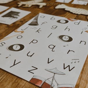 Pu&ograve; includere: Una carta bianca con l'alfabeto e illustrazioni di strumenti musicali. Le lettere sono in un font semplice e le illustrazioni includono una batteria, un clarinetto e note musicali. La carta &egrave; su una superficie di legno.