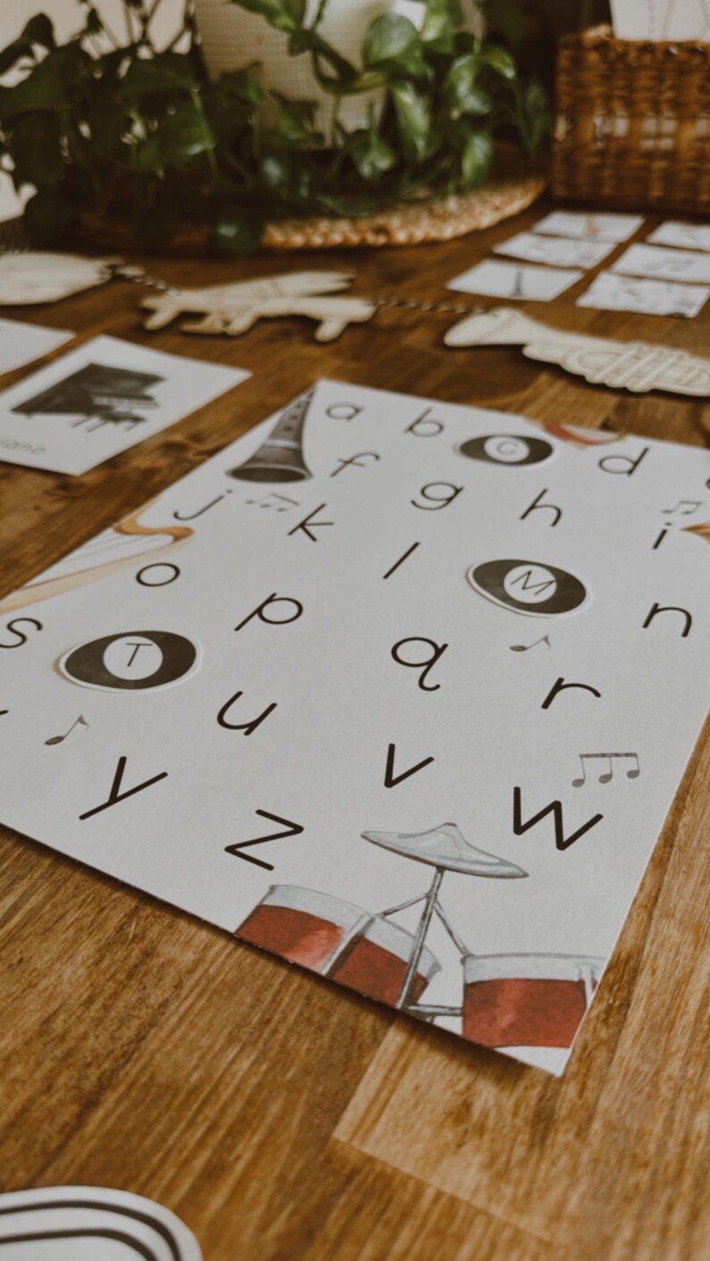Pu&ograve; includere: Una carta bianca con l'alfabeto e illustrazioni di strumenti musicali. Le lettere sono in un font semplice e le illustrazioni includono una batteria, un clarinetto e note musicali. La carta &egrave; su una superficie di legno.