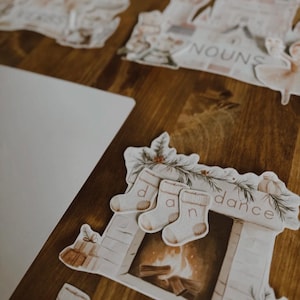 Peut inclure: Des d&eacute;coupes de papier d&eacute;coratives sur une surface en bois. Les d&eacute;coupes incluent une chemin&eacute;e avec des chaussettes &eacute;pelant "dance", un ch&acirc;teau et d'autres formes. La palette de couleurs est douce, avec des tons beige, blanc et marron.