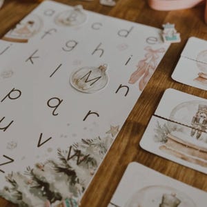 Peut inclure: Une table en bois pr&eacute;sente des cartes &eacute;ducatives avec l'alphabet, des illustrations sur le th&egrave;me de No&euml;l et des pi&egrave;ces de puzzle. Les cartes pr&eacute;sentent des lettres, un motif de sapin de No&euml;l et une boule &agrave; neige avec un casse-noisette. La palette de couleurs comprend le blanc, le vert et le marron.