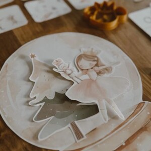 Peut inclure: Illustration fantaisiste d'une ballerine et d'un sapin de No&euml;l dans une boule &agrave; neige. La ballerine porte une robe rose et une couronne. Un petit soldat en jouet est &eacute;galement pr&eacute;sent. Le design est dans des tons aquarelle doux, avec une esth&eacute;tique chaleureuse.