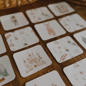 Peut inclure: Ensemble de cartes carr&eacute;es avec des illustrations &agrave; l'aquarelle d'un ch&acirc;teau, d'un casse-noisette, d'une ballerine et d'autres images sur le th&egrave;me des f&ecirc;tes. Les cartes sont dispos&eacute;es sur une surface en bois, avec un panier en osier en arri&egrave;re-plan.