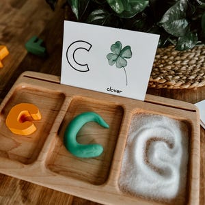 May include: A wooden tray with three compartments. The compartments contain a plastic letter C, a green playdough letter C, and a letter C drawn in white sand. A white card with a black letter C and a green clover with the word "clover" printed below is leaning against the tray.