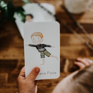 May include: A white card with a watercolor illustration of a child in a yoga pose. The child is wearing a black shirt and plaid shorts. The card reads "Plane Pose" in a simple font. The card is held in a child's hand.