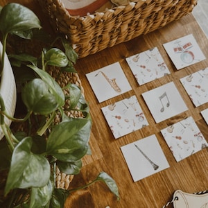May include: Wooden musical instrument cutouts and flashcards with musical instrument illustrations are arranged on a wooden surface. A woven basket holds additional wooden instruments. A plant is in the corner.