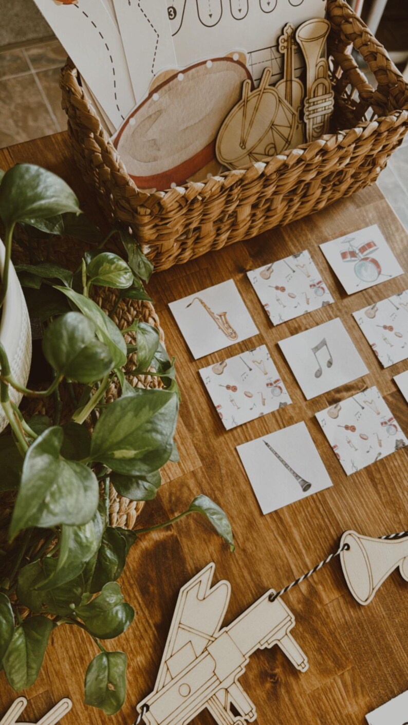 Pu&ograve; includere: Ritagli di strumenti musicali in legno e flashcard con illustrazioni di strumenti musicali sono disposti su una superficie di legno. Un cestino di vimini contiene altri strumenti in legno. Una pianta &egrave; nell'angolo.