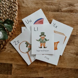 May include: A set of alphabet flash cards featuring illustrations of various objects. The cards are white with black lettering and colorful illustrations. The letters "LI" are shown with a leprechaun illustration, "UU" with a pink umbrella, "ZZ" with a magnifying glass, and "X" with a flag.