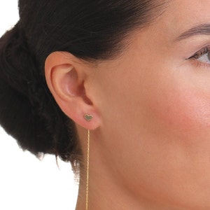 May include: A gold heart-shaped earring with a long chain dangling from it. The earring is worn in the left ear of a person with dark hair.