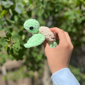 May include: A crocheted green and beige turtle toy with black eyes. The turtle is being held by a hand.