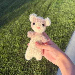 May include: A small, handmade teddy bear with a light beige body and light pink accents on the ears, paws, and nose. The bear has black eyes and is made of a fuzzy material. The bear is approximately 5 inches tall.