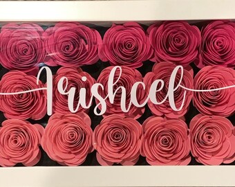 May include: A white frame with a glass front containing a display of pink paper flowers. The name "Trishcel" is written in white lettering on the glass.