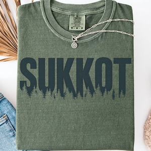 Może przedstawiać: Oliwkowa koszulka z napisem "SUKKOT" w ciemnoniebieskich literach nad sylwetką lasu. Srebrny naszyjnik z wisiorkiem w kształcie słońca spoczywa na koszulce. Koszulka jest złożona, z widocznymi dżinsami i butem.