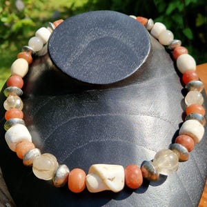 May include: A necklace with a mix of cream, orange, and clear beads, complemented by silver-coloured metal elements. The necklace is presented on a black mannequin, highlighting its design and colour variations. The beads are of various shapes and sizes, creating a unique aesthetic.