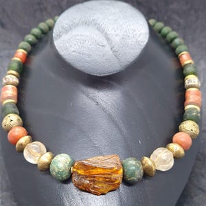 Könnte beinhalten: Eine Halskette mit einer Vielzahl von Perlen in Grün-, Orange-, Gold- und Klartönen. Der Mittelpunkt ist eine große, bernsteinfarbene, unregelmäßig geformte Perle. Die Halskette wird auf einer schwarzen Schaufensterpuppe präsentiert.