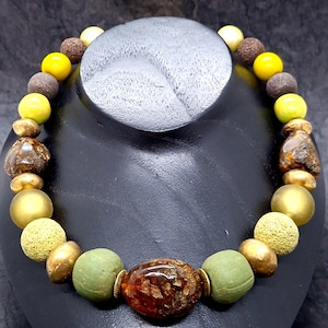 Könnte beinhalten: Eine Halskette aus grünen, gelben und braunen Perlen. Die Perlen sind in verschiedenen Größen und Formen, darunter rund, oval und unregelmäßig. Die Halskette wird auf einer schwarzen Puppe präsentiert.