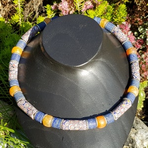 Op de afbeelding: Een blauwe en oranje ketting met witte en oranje gepatroneerde kralen. De ketting is gemaakt van glas en ligt op een zwarte displaystandaard.