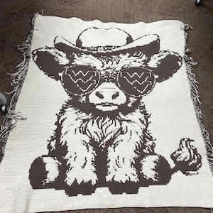 May include: A crocheted blanket featuring a brown and white cow wearing a cowboy hat and sunglasses. The cow is sitting down with its legs crossed. The blanket has a fringe border.