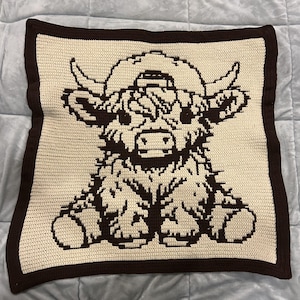 Pode incluir: Uma almofada de crochet castanho com um design de crochet branco de uma vaca das Terras Altas a usar um chapéu.