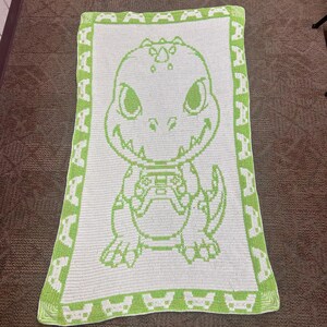 May include: A rectangular crochet blanket featuring a white center with a lime green graphic of a cartoon dinosaur holding a video game controller. The border is lime green with a repeating white pattern of small video game controllers.