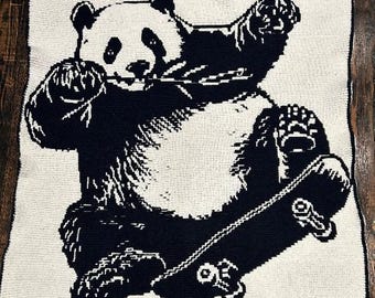 May include: A black and white crocheted blanket depicting a panda bear on a skateboard. The panda is in a dynamic pose, with its arms raised and legs bent. The blanket has a black border.
