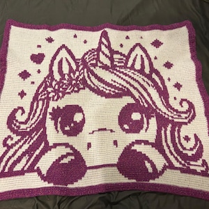 May include: A crocheted blanket featuring a purple and white design of a cartoon unicorn. The unicorn has large eyes, a floral crown, and flowing hair. The blanket has a scalloped purple border and decorative star and heart accents.
