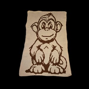 May include: A crocheted blanket featuring a smiling monkey design in brown against a cream background. The monkey is centered on the blanket, with its arms and legs visible. The blanket has a rectangular shape and a textured appearance.