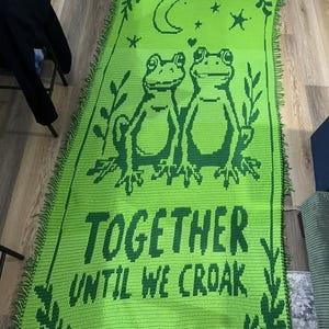 May include: A green crocheted blanket with a design of two frogs, a crescent moon, stars, and the text "TOGETHER UNTIL WE CROAK." The blanket has a fringed edge and a dark green border with decorative elements.