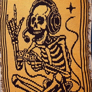 May include: Mustard yellow and black crocheted blanket featuring a skeleton wearing headphones and playing a video game. The blanket has the text "THE GAMER" and other symbols. The blanket is rectangular with a fringed edge.