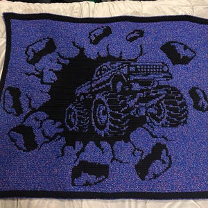 May include: A blue and black crocheted blanket with a black monster truck design breaking through a wall. The blanket has a black border and is made of a soft, textured yarn. The design includes black rock fragments.