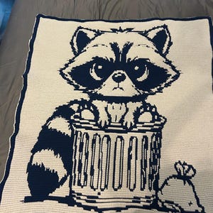 May include: A cream-colored, crocheted blanket featuring a navy blue raccoon design. The raccoon is depicted peeking out of a navy blue trash can, with a navy blue trash bag beside it. The blanket has a navy blue border.
