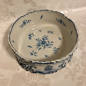 Puede incluir: Un cuenco de cerámica blanca con un diseño floral azul y blanco. El cuenco tiene un borde festoneado y un diseño en relieve alrededor de la base.