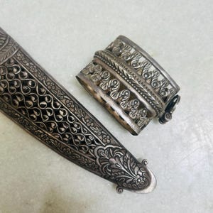 May include: A silver ornate dagger sheath with intricate floral designs. The sheath is made of metal and has a decorative, openwork design.