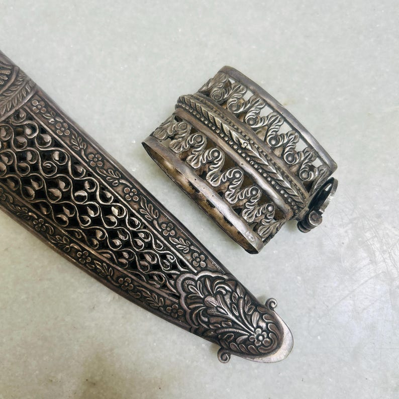 May include: A silver ornate dagger sheath with intricate floral designs. The sheath is made of metal and has a decorative, openwork design.