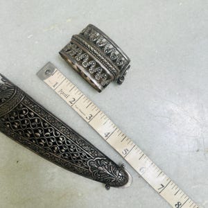 May include: A silver dagger with intricate floral designs and a silver decorative piece with a similar design. The dagger is 6 inches long and the decorative piece is 2 inches long.
