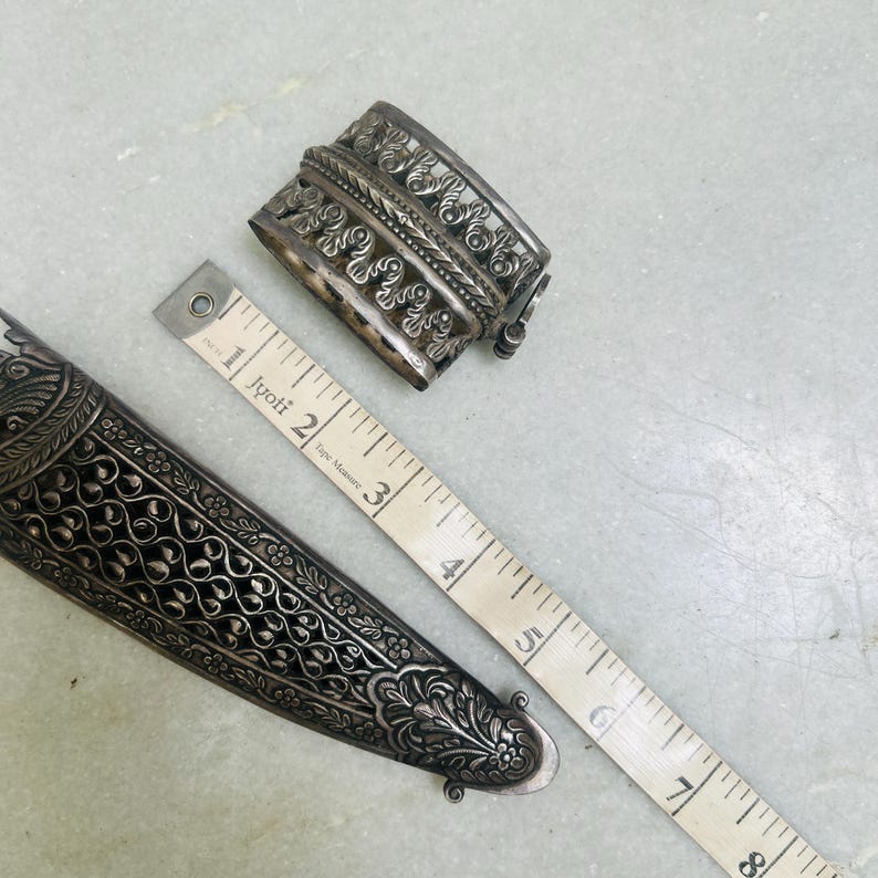 May include: A silver dagger with intricate floral designs and a silver decorative piece with a similar design. The dagger is 6 inches long and the decorative piece is 2 inches long.