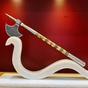 Peut inclure: Une hache ornée avec une tête argentée et un manche en bois à motifs. La tête de la hache est gris foncé et le manche présente un motif géométrique répété en vert et jaune. La hache repose sur une sculpture blanche et incurvée sur fond rouge.