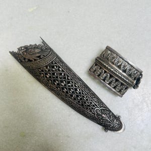 May include: Two silver ornate pieces of jewelry, one is a long, narrow piece with intricate floral and bird designs, the other is a shorter, wider piece with a more geometric design.