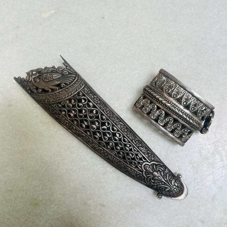 May include: Two silver ornate pieces of jewelry, one is a long, narrow piece with intricate floral and bird designs, the other is a shorter, wider piece with a more geometric design.