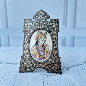 May include: A silver picture frame with intricate floral designs and a painted portrait of a woman wearing a colorful headdress and jewelry.