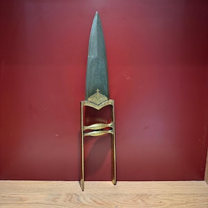 May include: An antique dagger with a long, pointed blade and ornate gold-colored handle. The blade has etched designs, and the handle features intricate patterns. The dagger is displayed against a red background, showcasing its historical and decorative qualities.
