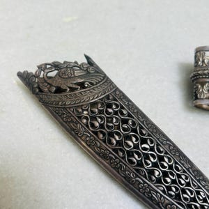 May include: A silver dagger sheath with intricate floral and peacock designs. The sheath is made of metal and has a detailed, ornate pattern.