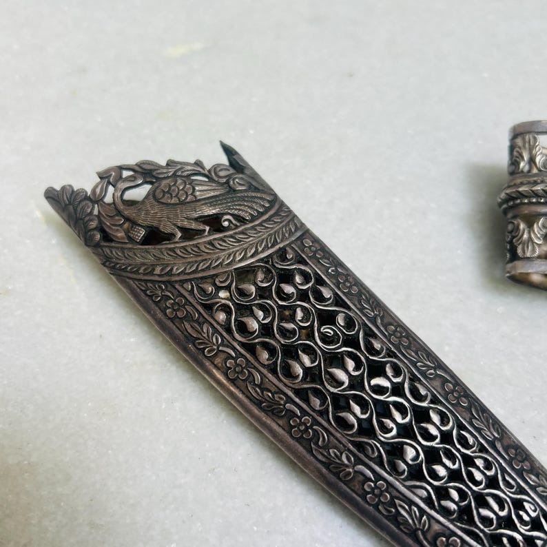 May include: A silver dagger sheath with intricate floral and peacock designs. The sheath is made of metal and has a detailed, ornate pattern.