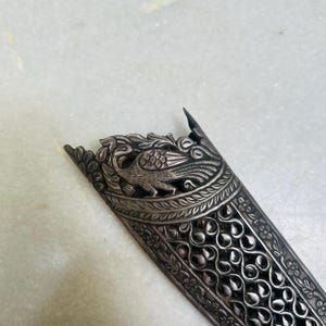 May include: A silver-colored dagger sheath with intricate floral and peacock designs. The sheath is made of metal and has a detailed, ornate pattern.