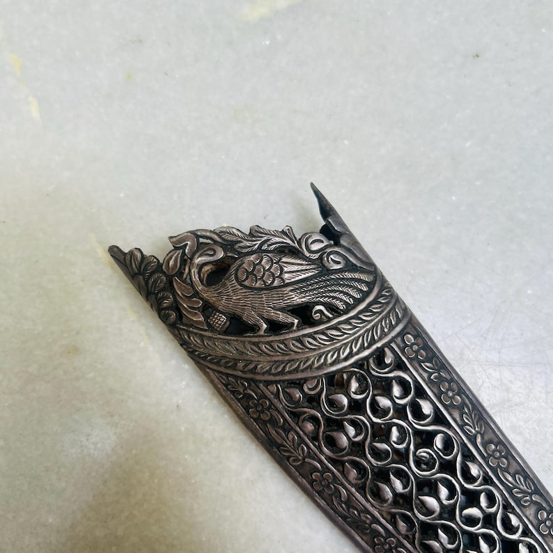 May include: A silver-colored dagger sheath with intricate floral and peacock designs. The sheath is made of metal and has a detailed, ornate pattern.