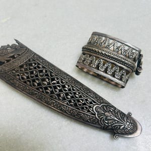 May include: A silver ornate dagger sheath with intricate floral and geometric designs. The sheath is paired with a silver decorative ring with similar designs.