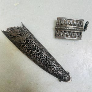 May include: Two silver ornate pieces, one is a long, narrow, decorative sheath with a bird design, the other is a shorter, wider, decorative cuff.