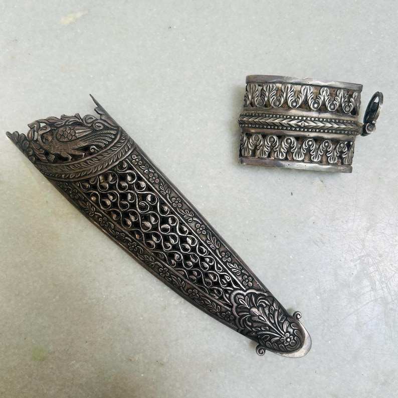 May include: Two silver ornate pieces, one is a long, narrow, decorative sheath with a bird design, the other is a shorter, wider, decorative cuff.