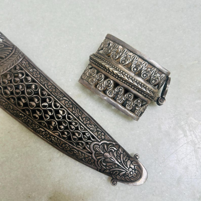 May include: A silver ornate dagger sheath with intricate floral designs. The sheath is closed with a silver clasp with a similar design.