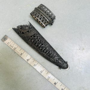 May include: A silver dagger sheath with intricate floral and bird designs. The sheath is approximately 7 inches long and has a decorative silver cap.